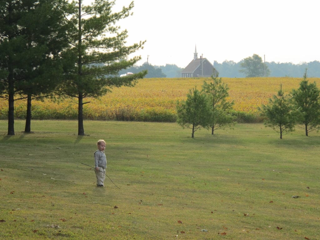 A Picturesque Indiana Setting with Our Son Playing with a Stick with farm land and a church behind him.